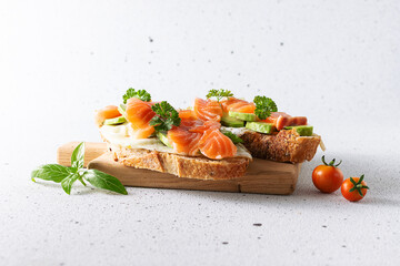 Tasty and healthy breakfast idea. Homemade sandwiches with salmon, eggs, avocado, parsley and full grain bread on light stone background close up.