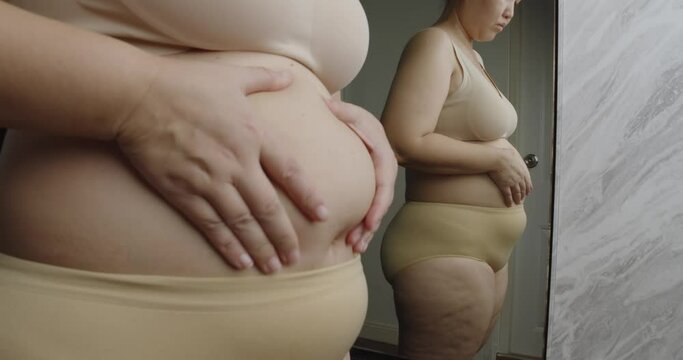 Asian oversize woman in front of a mirror looks at her body. Excess belly fat, Unhealthy lifestyle, Diet concept.