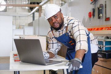Black male carpenter using professional laptop in furniture carpentry room New design concept in workshop