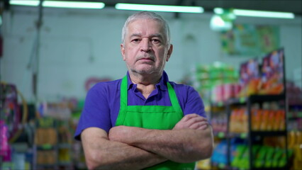 One serious older employee of grocery store crossing arms with stern worried expression stands inside supermarket local shop