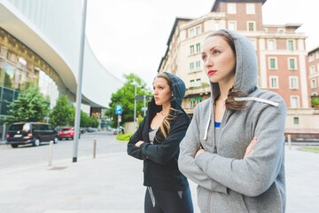 Two young beautiful millennial sportive woman wearing tracksuit outdoor in the city, arms crossed, overlooking - sportive, health, training concept