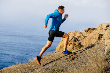 athlete runner running mountain uphill in blue jacket and black tights, background of sea