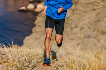 front view runner running mountain uphill in blue jacket and black tights, trail dry grass