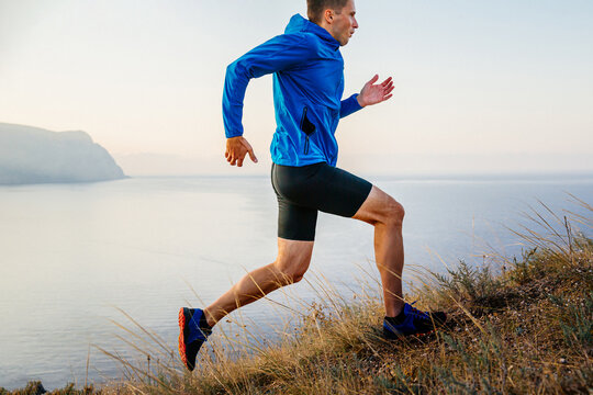 male runner athlete running uphill in blue jacket and black tights, background of sky and sea
