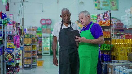 Two senior diverse employees working at supermarket looking at tablet device, teamwork scene of...