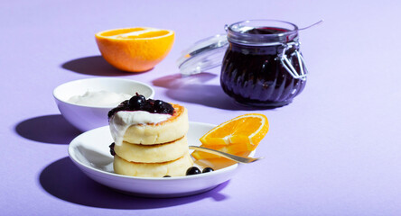 Breakfast of syrniki with sour cream and blackcurrant jam with orange on a lilac background.