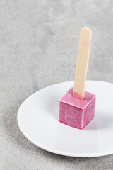 Mini Black currant ice cream in the shape of a cube on a wooden stick. On a plate. Light gray background. Copy space