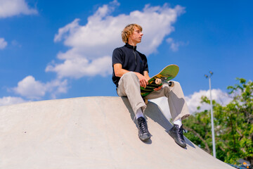 A young guy skateboarder with long hair sits on his skateboard on the edge of a skate pool at a...
