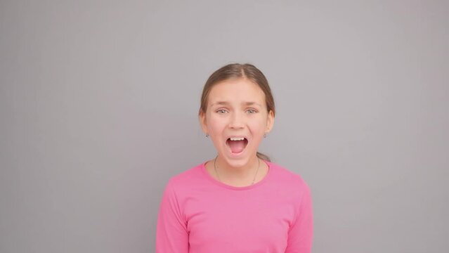 A ten year old girl looks at the camera and screams in a pink t-shirt on a gray background.
