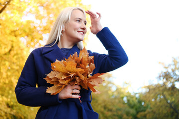 Beautiful young blonde woman with long hair, in a blue coat and with a bouquet of autumn leaves in her hands, smiles and poses in an autumn park