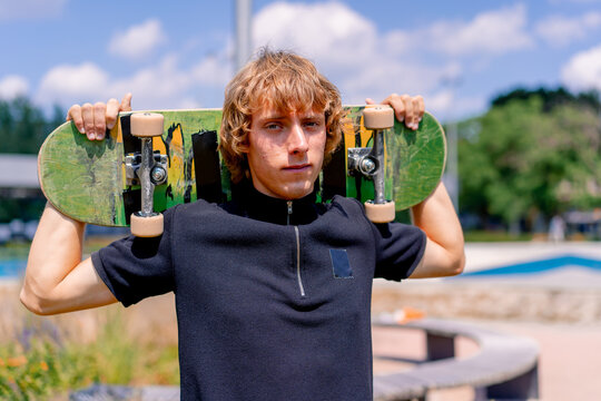 Portrait of a young serious guy skater with long hair holding a skateboard on his shoulders on the background of skatepark and sky