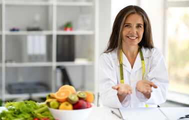 Happy european middle aged lady doctor nutritionist hold free space at table with fruits, vegetables