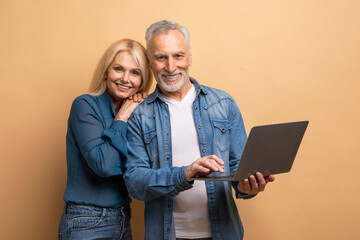 Loving elderly spouses using computer laptop on beige background