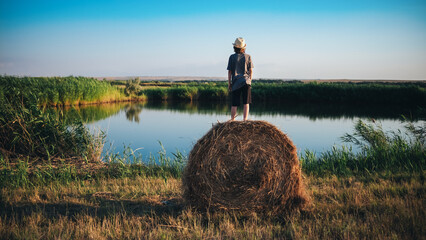 A young man in a hat stands on a haystack and looks at the surface of the lake in the reeds