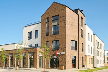 Part of large street with corner of long three storey brick block of flats with cafe on facade standing by low building with glass doors