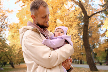A young dad holds a newborn daughter in purple overalls in his arms. Dad walks with a child in the autumn park