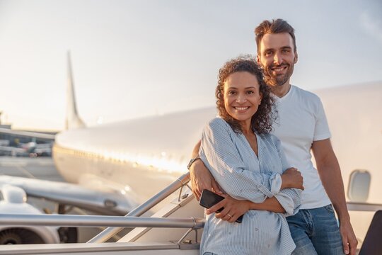 Portrait of happy couple of tourists, man and woman looking excited while standing together outdoors ready for boarding the plane at sunset. Vacation, lifestyle, traveling concept - Powered by Adobe