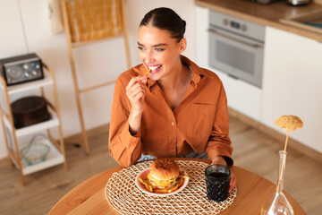 Cheerful young woman holding and eating french fries and hamburger, sitting at table in kitchen...