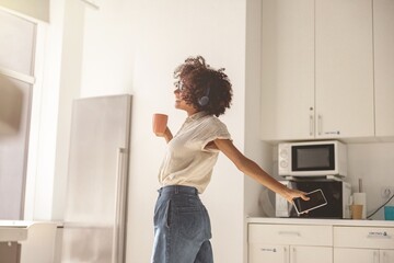 Fototapeta na wymiar Happy African American lady wearing wireless headphones and dancing in the kitchen while holding the mobile phone