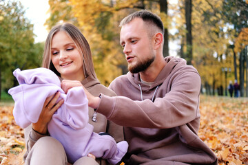 Young parents sit on a plaid with a newborn baby in an autumn park. The mother holds the child in her arms, the parents look tenderly at their daughter