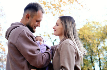 Fototapeta premium Young mother and father are walking with a newborn baby in the autumn park. The father looks at the child with tenderness, the wife looks at the husband with love