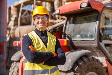 Cheerful matured man in safety helmet and work vest looking at camera and smiling while keeping arms crossed