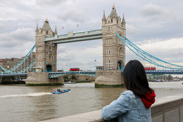 Female Tourist in London, looking at famous and iconic sights of the city as red buses cross the Tower Bridge, on the banks of the River Thames