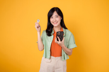 future of finance with cheerful young Asian woman in her 30s, donning orange shirt and green jumper, displaying smartphone screen and crypto currency coin on yellow background. Future finance concept.