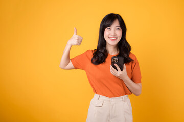 Experience happiness with a young Asian woman in her 30s, donning an orange shirt, showing thumb up gesture while using smartphone on yellow background.