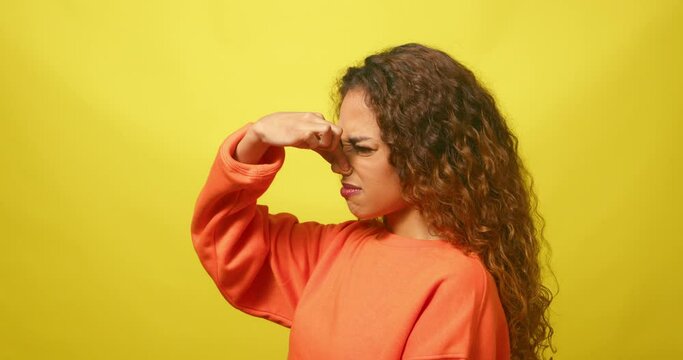 Beautiful young woman pinches nose smells bad odour stink, yellow studio