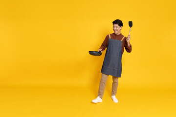 Young Asian man chef wearing kitchen apron cooking and holding pan and spatula isolated on yellow background, Full body composition