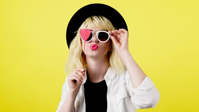 Wink, flirt and hipster woman sending air kiss and happy excited smiling. Fashionable lady blowing a kiss with confidence flirty emotion for cosmetics makeup, trendy outfit in studio yellow background