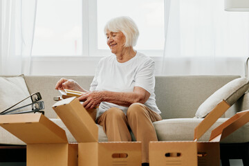 elderly woman sits on a sofa at home with boxes. collecting things with memories albums with photos and photo frames moving to a new place cleaning things and a happy smile. Lifestyle retirement. - obrazy, fototapety, plakaty