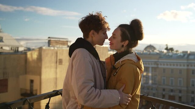 4k Lgbt couple in love kisses and hugs on rooftop in city spbd. Closeup of young woman kissing with tenderness and hugging, looking with smiles and standing on background of the town outdoors