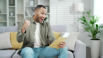 Happy young man receives letter with good news notification with great result sitting at home on sofa. indoor. Male getting offer win won winning victory triumph congratulations feeling happiness joy
