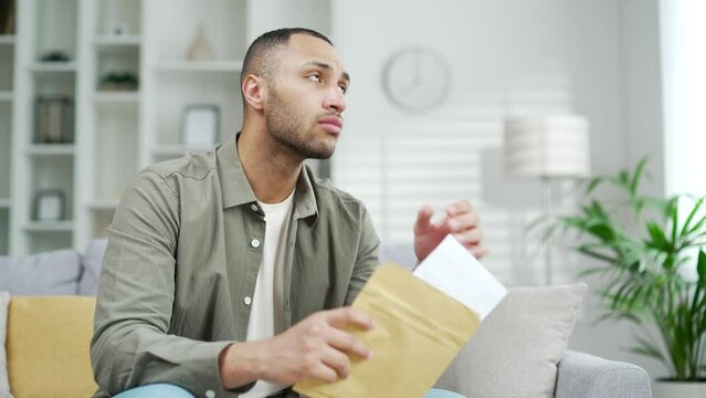 Unhappy young man opening envelope with letter about financial problem bank debt bill medical result sitting on couch in living room at home Upset male received bad new by mail student failure refusal