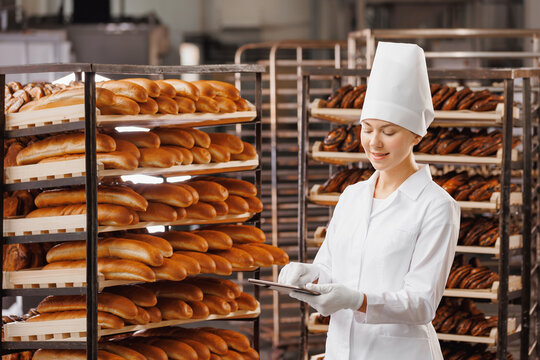 Baker woman in chef uniform use tablet computer for control quality of craft bread in bakery factory, sun light. Banner modern food industry