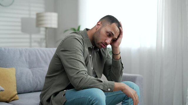 Silhouette of a young adult man suffering from depression sitting at home on the couch. The male has mental health issues upset guy alone near a window One anxious depressed man in despair indoor