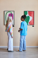 Vertical full length portrait of two teen girls discussing pictures in modern art gallery