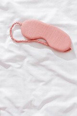 Sleeping mask pink color on bed white bedclothes background. Eye cover for best sleep. Concept of home comfort and wellbeing, dream well, comfort rest at night. Top view sleep mask, copyspace