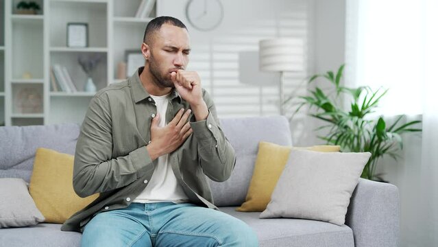 A sick man coughs holding his chest while sitting on the sofa in the living room at home. An unhealthy mixed race black male with inflammation from a cold or flu coughs indoors