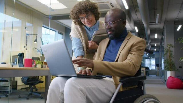 Low angle of male African American employee in wheelchair showing project ideas to female Caucasian coworker indoor at daytimeLow angle of male African American employee in wheelchair showing project 