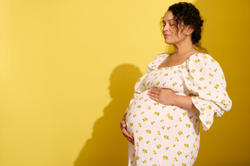 Happy gravid pregnant woman gently stroking and hugging her big belly, isolated over yellow studio...