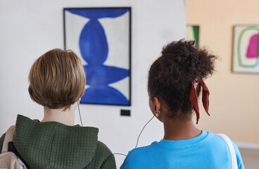Back view closeup of two teenagers looking at pictures in modern art gallery or museum sharing earphones with audio guide