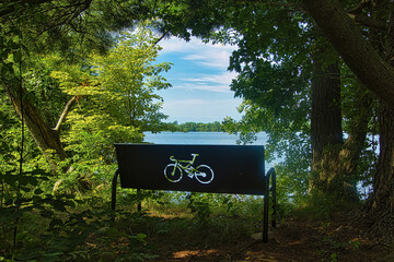 On a sunny Summer day, an empty bench with a bicycle design faces the Chippewa River from a shady spot along the Old Abe State Trail, near Jim Falls, WI. - obrazy, fototapety, plakaty