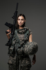 Beautiful female soldier with stunning blue eyes stands in her military uniform holding an automatic rifle for a studio portrait taken against a grey background.