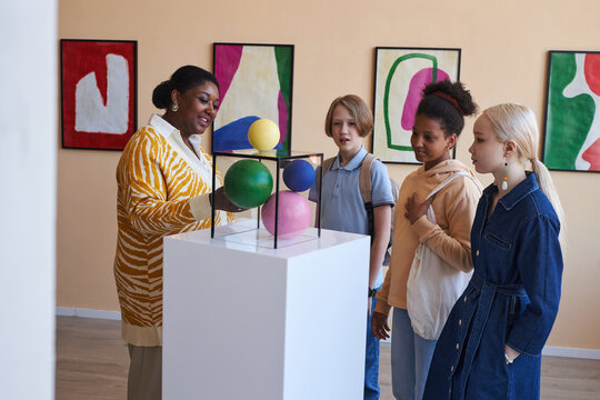 Diverse group of teenagers listening to teacher or tour guide in modern art gallery or museum
