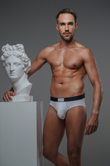 Fototapeta na wymiar A stunning male model with a chiseled torso and in underwear striking a pose next to a bust of an ancient Greek sculpture