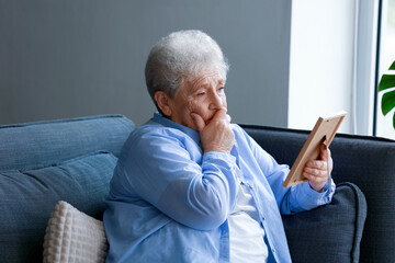Sad senior woman with photo frame sitting on sofa at home