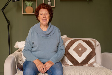 Portrait of a modern elderly woman with blue eyes and red hair sits on a sofa and looks at the camera